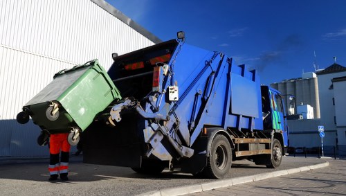 A large blue waste collection truck with an open rear loading area positioned on a paved street in an urban area, likely in Hammersmith, with a clear blue sky overhead. The truck has a hydraulic lifting mechanism at the back, actively raising and tipping a green wheeled waste bin for emptying. The green bin, made of rigid plastic, has two black wheels at the base and a black handle on the lid, which is open during the waste disposal process. Partially visible behind the truck, a worker wearing high-visibility red and dark clothing with reflective strips stands near the waste bin, overseeing the loading. In the background, a white industrial or commercial building with vertical ribbed siding is evident, along with other urban infrastructure. The scene captures the routine activity of rubbish removal, consistent with the services offered by Office Clearance Hammersmith, demonstrating the transportation and handling of waste materials in a professional urban setting.