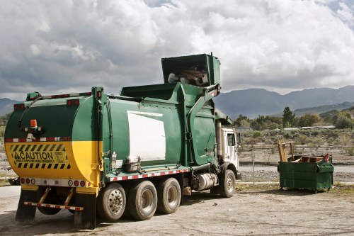 A green waste collection truck with a cylindrical rear compactor, parked on a dirt surface with a partly cloudy sky overhead. The truck features yellow caution markings along the lower rear section and appears to be in the process of collecting rubbish. To the right of the vehicle, there is a green open-top skip filled with assorted waste materials, positioned on the same dirt ground. In the background, a chain-link fence separates the area from a landscape of distant hills and sparse vegetation, suggesting a semi-rural or industrial setting. The overall scene indicates a rubbish removal service operation, potentially serving the Hammersmith area, with a focus on waste collection and disposal activities.