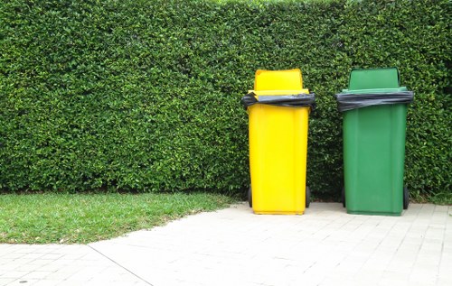 Two large plastic wheelie bins, one yellow and one green, are positioned on a paved sidewalk in front of a neatly trimmed hedge. The yellow bin is slightly taller and has a black lid that is partially open, with some dark rubbish visible inside. The green bin, which is slightly shorter, has a closed lid. The surface underneath the bins consists of light grey paving stones, while the dense hedge behind them provides a natural backdrop. The scene is well-lit with natural daylight, emphasizing the vibrant colours of the bins against the lush green foliage, illustrating a typical urban or suburban rubbish disposal area consistent with rubbish removal services in the Hammersmith area, as managed by Office Clearance Hammersmith.