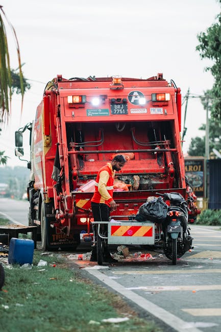 A worker wearing a yellow high-visibility vest with red reflective strips is operating a red rubbish collection truck parked on the side of a street, performing waste disposal. The truck's rear is ope