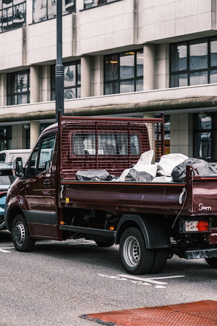 A dark red flatbed pickup truck is parked on an urban street in front of a modern office building with large glass windows and a curved concrete facade. The truck's bed is loaded with various types of