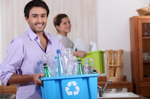 In a domestic kitchen setting, a young man with dark hair and a beard, dressed in a light purple shirt, is smiling and holding a blue recycling bin filled with various glass bottles and jars, including green and transparent types, with visible textures of glass reflecting light. Behind him, a woman with dark hair tied back, wearing a light-colored top, stands by a countertop with a green plastic storage container and some stacked white items, possibly kitchenware or packaging. The background features wooden kitchen furniture, including a cabinet and a shelf, with neutral cream curtains covering the windows, indicating daytime lighting. The scene suggests an environment of waste sorting or rubbish collection preparation aligned with professional rubbish removal services like those provided by Office Clearance Hammersmith, situated in or near London postal districts, with a focus on responsible waste management and recycling practices.