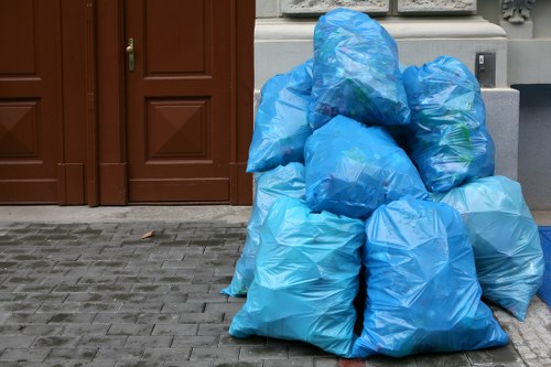 A collection of several large blue plastic rubbish bags filled with waste, stacked against a brown wooden door on a paved pavement. The bags are tightly sealed, with some showing folds and creases in the plastic material. They occupy most of the foreground, positioned directly on the gray stone surface, which appears to be part of a driveway or pavement area outside a building. Behind the bags, there is a section of a gray wall and a small part of an electrical outlet visible to the right. This scene illustrates typical waste collection or rubbish removal activity, consistent with services offered by Office Clearance Hammersmith in the local Hammersmith area, specifically in the postal district W6, where domestic or small commercial waste might be gathered for collection or disposal.